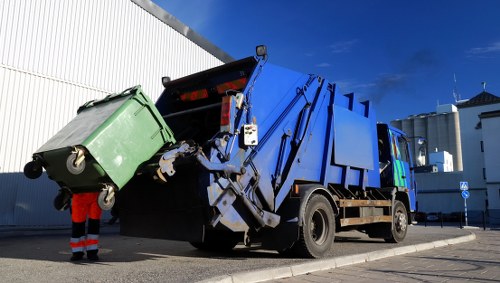 The image depicts a blue and black rubbish collection truck with an open top, equipped with a hydraulic lifting mechanism at the rear, actively emptying a green wheeled waste bin into its hopper. The green bin, made of plastic, is positioned at the back of the truck, with its lid open to allow contents to spill into the vehicle. The truck is parked on a paved surface near a large white industrial building or warehouse, with a clear blue sky overhead, indicating bright sunlight. A worker dressed in an orange high-visibility jacket and dark trousers stands nearby, guiding or overseeing the collection process. The scene suggests a routine rubbish removal operation typical for commercial or municipal waste services, such as those provided by Tower Hamlets House Clearance, and highlights standard equipment used for efficient waste collection in urban areas of Tower Hamlets or surrounding London boroughs. The image emphasizes functionality, safety gear, and the logistics involved in rubbish disposal services within the local environment.