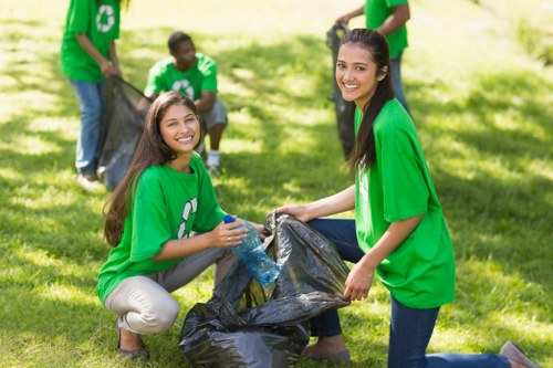 A group of young volunteers participating in a community litter pick in a grassy outdoor area, wearing matching bright green T-shirts with a white recycling symbol on the front. Two women in the foreground are smiling and kneeling on the grass while holding a large black rubbish bag open, one placing a plastic bottle into it. In the background, two more volunteers are seen holding another rubbish bag, actively collecting trash from the ground, with trees and sunshine creating a pleasant, well-lit environment. The scene depicts an organized outdoor cleanup activity, highlighting community engagement in waste management and environmental preservation, consistent with the efforts of local rubbish removal services such as Tower Hamlets House Clearance, operating in East London areas.