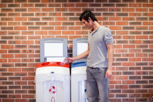 A young man with dark hair, dressed in a light grey long-sleeved shirt and grey trousers, is seen standing outdoors beside two large recycling bins with white bodies and grey lids, positioned against a red brick wall. He is holding a small plastic bottle with clear water, preparing to dispose of it in the recycling bin on the left, which features a red recycling symbol on the front. The scene takes place on a paved surface, possibly a street or driveway, with the bins situated close to the brick wall, indicating a typical setting for rubbish collection or waste disposal services in Tower Hamlets. The lighting appears natural, and the overall scene reflects a routine rubbish or recycling task with the presence of commercial or residential waste management mechanisms from Tower Hamlets House Clearance, illustrating environmentally conscious waste separation practices.